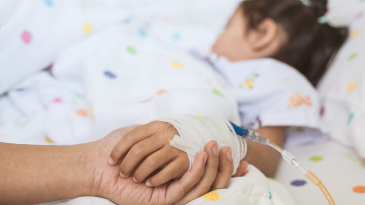 Mother hand holding sick daughter hand who have IV solution bandaged with love and care while she is sleeping on bed in the hospital