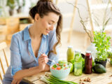 Young and happy woman eating healthy salad sitting on the table with green fresh ingredients indoors